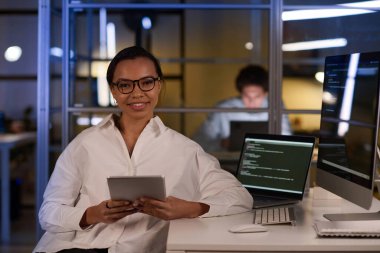 Elinde dijital tablet bulunan gözlüklü genç bir kadının orta boy portresi akşam geç saatlerde iş yerinde oturuyor.