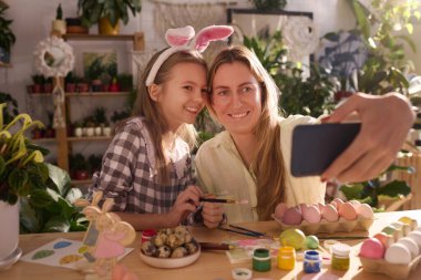 Paskalya için yumurta boyarken akıllı telefondan selfie çeken yaratıcı anne ve kızın orta boy portresi.