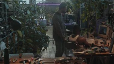 Giyinik bir erkek, arkadaşıyla konuşuyor bilgisayardan CCTV izliyor, sonra kıyamet sonrası sığınakta yaşarken tornavidayla lambayı tamir ediyor.