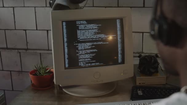 ポスト黙示録のバンカーでデスクの古いコンピュータでコーディングするヘッドフォンのブラックプログラマーの肩のショットに — ストック動画