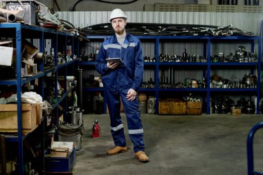 Dijital tablet taşıyan beyaz üniformalı genç bir adamın fabrika deposunda fotokopi çekerken görüntüsü.