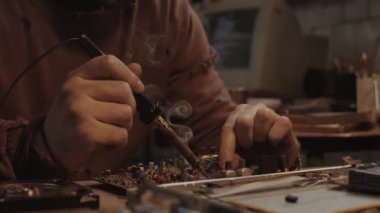 Mahşer sonrası sığınaktaki masada elektronik parçaları tamir ederken lehimleme demiri kullanan yıpranmış bir adamın görüntüsünü kapat