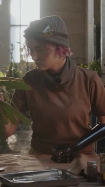 Protez kollu bir kızın dikey portresi yeşil bitkilerle ilgileniyor ve kıyamet sonrası sığınaktaki kameraya bakıyor.