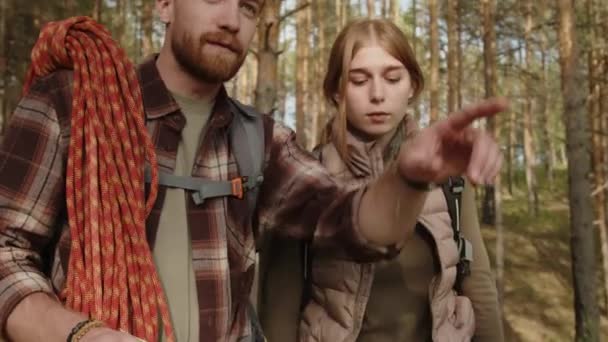 松の森で一緒に立っている間 紙の地図上のハイキングルートを議論している男性と女性のハイカーの若いカップル — ストック動画