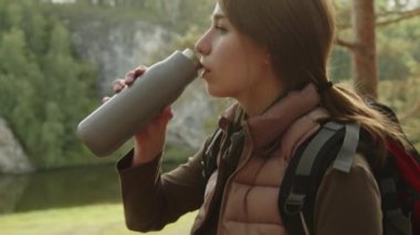 Genç bayan yürüyüşçünün yaz günü doğada dolaşırken ormanda dikilip şişeden su içişini çek.