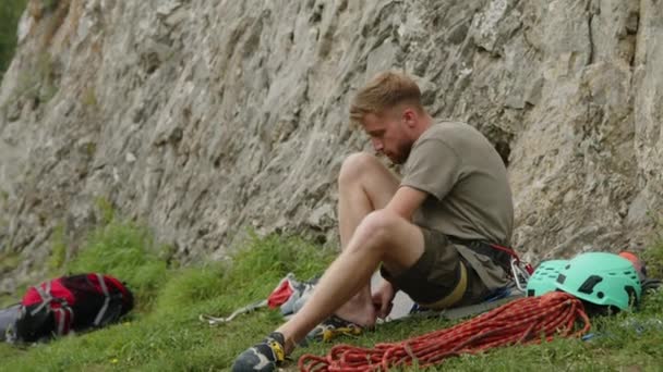 Mannelijke Bergbeklimmer Zittend Mat Aan Voet Van Rots Het Aantrekken — Stockvideo