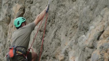 Miğferli ve koşumlu genç bir adam halatla kayaya tırmanıyor, kısa bir mola veriyor ve eline tebeşir alıyor.