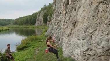 Genç bir kadın halatla kayadan iniyor, erkek arkadaşına beşlik çakıyor ve nehir kıyısına tırmanmaktan bahsediyor.