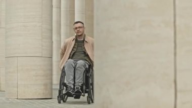 Günlük giysili ve gözlüklü genç adam şehir merkezindeki sütunlar boyunca tekerlekli sandalyeyle kameraya doğru gidiyor.
