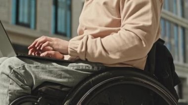 Tekerlekli sandalyede oturan ve gün boyunca dışarıda dizüstü bilgisayarda çalışan tanınmamış erkek serbest çalışanın yakın plan çekimleri.