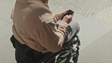 Yüksek açılı, engelli bir adamın tekerlekli sandalyede dışarıda otururken ve akıllı telefon ekranında gezerken gün boyu mobil internet kullanırken görüntüsü.