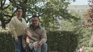 Orta uzunlukta tekerlekli sandalyedeki genç adam ve kız arkadaşının parkta gülümseyerek kameraya poz verişi.