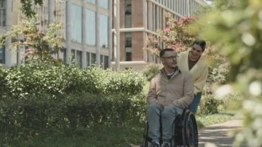 Parkta yürürken tekerlekli sandalyeye mahkum eden genç adamla gülümseyen ve sohbet eden bir kadın.