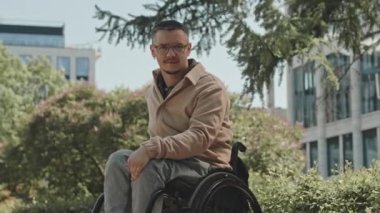 Parkta güneşli bir günde tekerlekli sandalyede kameraya poz veren genç bir adam.