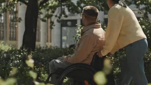 Śledzenie Ujęcia Kobiety Pchającej Mężczyznę Wózku Inwalidzkim Przez Park Rozmawiającej — Wideo stockowe