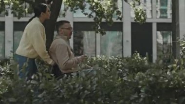 Güneşli bir günde parkta yürürken arkadaşını tekerlekli sandalyeyle iten ve onunla konuşan kadının yan görüntüsü.