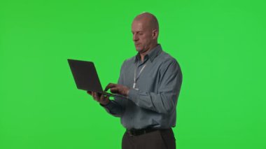 Orta yaşlı veri merkezi teknisyeninin yeşil krom anahtar duvarına karşı duran ve dizüstü bilgisayar üzerinde çalışan stüdyo görüntüsü.