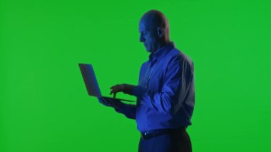 Veri merkezi uzmanı mavi neon ışığın altında duruyor, elinde dizüstü bilgisayar ve yeşil krom anahtar arka planlı daktilo kodu var.