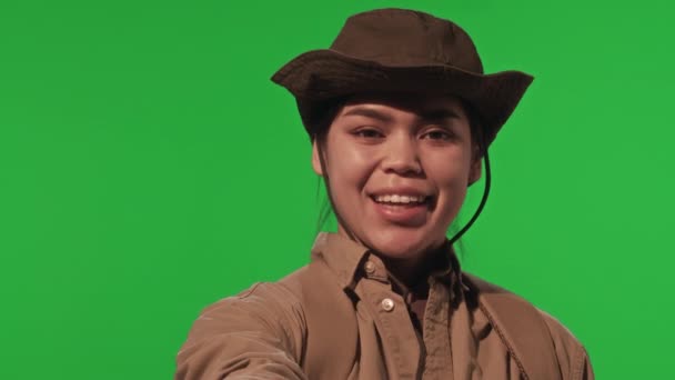 穿着卡其布衣服 头戴帽子的年轻而快乐的女性游客 穿着卡其布衣服 在镜头前微笑着 并在绿色的彩色背景上显示出V字迹 — 图库视频影像