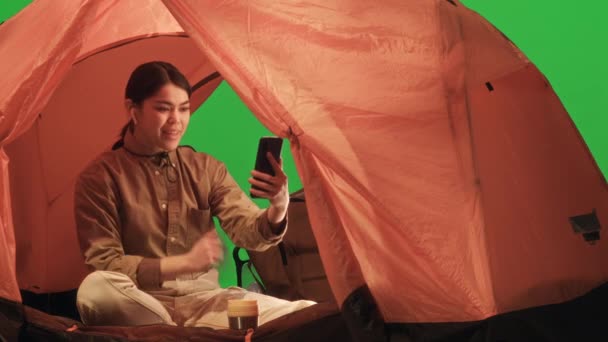 年轻的女游客坐在绿色色彩艳丽的帐篷里 一边微笑着一边挥手 一边用手机在大自然中的远行中打电话 — 图库视频影像