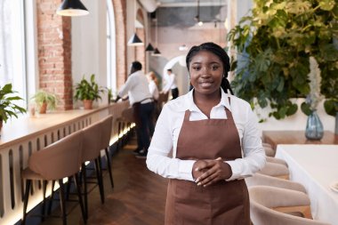 Beyaz gömlek ve kahverengi önlükle modern restoranda kameraya poz veren orta boy Afrikalı Amerikalı garsonun portresi.