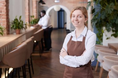 Beyaz tişörtlü, kahverengi önlüklü, kendine güvenen beyaz bir garsonun orta boy portresi. Kameraya gülümsüyor.