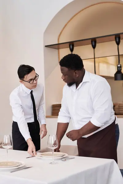 Vertical medium long shot of Asian headwaiter and African American busser discussing work while setting table in modern restaurant