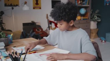 Küçük kıvırcık saçlı Afrikalı Amerikalı çocuk çocuk çocuk odasında oturuyor ve renkli kalemlerle kağıda resim çiziyor.