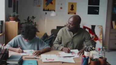 Afrikalı Amerikalı baba ve küçük oğlu oturma odasında oturmuş, renkli kalemler kullanıyor ve evde boş zamanlarında birlikte resim çizerken konuşuyorlar.