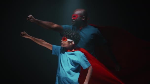 アフリカ系アメリカ人の父親と小さな息子は 赤いマスクを着て腕を抱え 暗い背景を持つスタジオでスーパーヒーローを飛ばすようなポーズをしました — ストック動画