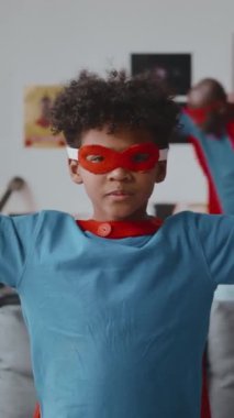 Süper kahraman pelerinli ve maskeli küçük kıvırcık saçlı Afro-Amerikan çocuğun dikey portresi. Babam oturma odasında koşarken bisepsleri ve poz veriyor.