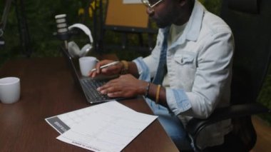 Genç Black Podcaster ses stüdyosunda kayıt bölümüne hazırlanırken dizüstü bilgisayarda çalışıyor ve notlar yazıyor.