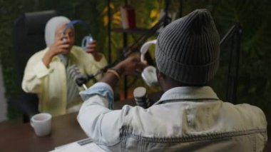 Erkek podcast sunucusu, kayıt stüdyosundaki bayan konukla konuşurken kulaklık takıyor ve mikrofonla konuşuyor.