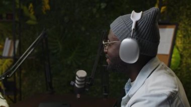 Genç Afrikalı Amerikalı podcast sunucusu mikrofonla gülümserken stüdyoda yeşil duvarlı bir röportaj kaydediliyor.