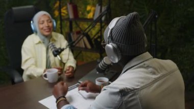Siyah podcast sunucusu mikrofonda konuşurken tesettürde gülümseyen ve stüdyoda çay içen genç bir kadınla röportaj yapıyor.