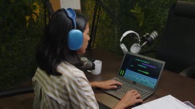 Kulaklıklı bir kadın kayıt stüdyosunda post prodüksiyon yaparken dizüstü bilgisayarla ses podcast bölümünü düzenliyor.
