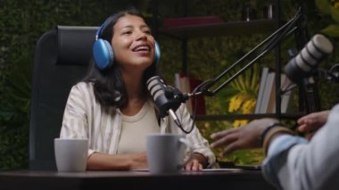 Genç Latin kadın blogcu kulaklıkla mikrofonla gülümsüyor ve mikrofonla konuşurken ses stüdyosunda yardımcı sunucuyla podcast yapıyor.