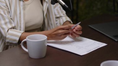 Latin kadın podcast sunucusunun kağıt plan ve çay fincanı ile masada otururken, elinde kalem tutarken ve stüdyoda yeni bölümler kaydederken mikrofona konuşurken yakın çekim görüntüsü.