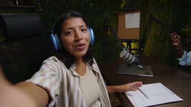 Kulaklıklı genç Latin kadının POV 'u kameraya bakıyor, gülümsüyor ve sosyal medya videolarını podcast stüdyosunda çekerken Afrikalı Amerikalı konukları anlatıyor.