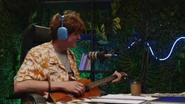 Genç kıvırcık saçlı, kulaklıklı, gitar çalan ve mikrofonla şarkı söyleyen yeşil duvarlı ve neon ışıklı kayıt stüdyosu tasarımlı bir müzisyen.