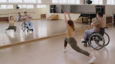 Tekerlekli sandalyedeki genç adam stüdyodaki ayna duvarının önünde kadın dans öğretmeniyle esneme egzersizleri yapıyor.