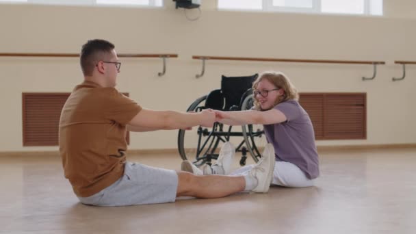年轻的残疾妇女面对男教练坐在地板上 坐在前弯 练习舞伴伸展身体 轮椅作为背景 — 图库视频影像