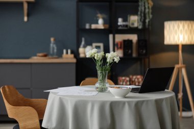 Seçici odak, cam vazodaki taze çiçeklerden, laptoptan ve modern apartmanın içindeki yemek masasındaki belgelerden çekilen insanlar değil.