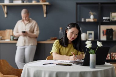 Genç Asyalı üniversite öğrencisi evde yemek masasında oturmuş çevrimiçi ders izliyor ve not alıyor. Kız arkadaşı arka planda duruyor.