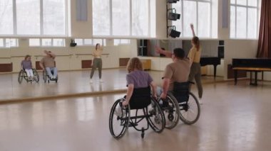 Tekerlekli sandalyedeki çift stüdyoda koreografi dersi sırasında kadın öğretmenle dans hareketleri yapıyor.