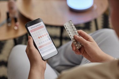 Doğum kontrol haplarıyla su toplayan ve akıllı telefondan takvimi kontrol eden tanınmayan genç bir kadının yüksek açılı görüntüsü.