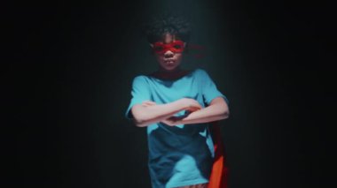 Süper kahraman gibi giyinmiş, kollarını kavuşturmuş, kameraya dönüp karanlık stüdyoda poz veren kendinden emin Afrikalı Amerikalı küçük çocuk.
