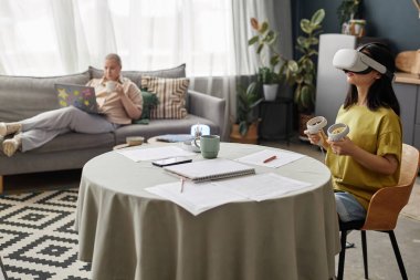 Kafkasyalı kız arkadaşı dizüstü bilgisayarla kanepede dinlenirken Asyalı kız öğrencinin VR kulaklık kullanmasının seçici odak noktası.