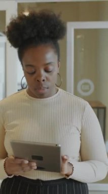 Genç siyahi iş kadınının dijital tablet kullanırken ve iş arkadaşını karşılarken ofiste yürürken dikey çekimleri.