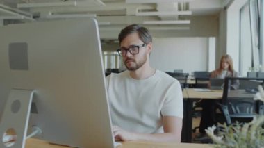 Genç iş adamı, modern ofis bilgisayarlarında internette sörf yapıyor, insanlar arka planda çalışıyor.